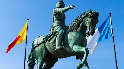 Joan of Arc bronze statue in Orleans, France. Joan is featured with sword drawn on a horse in battle dress, flanked by a blue and white flag plus a red and yellow flag