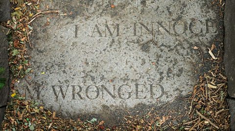 An inscribed gravestone flat on the ground reads I am innocent, I am wronged. Leaves and twigs cover the corners, part of the Salem Witch Trial memorial