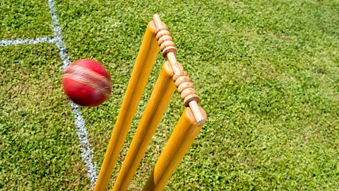 A red cricket ball approaches a set of wooden stumps and bails at speed. The surface is very green, short grass