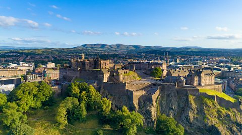 An aerial view of Edinburgh Castle on a sunny day, with greenery and trees at the forefront and the city centre at the back