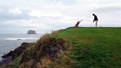 A man playing golf by sea in Aberlady Bay, Scotland, on a cloudy day. He stands on the grass which overlooks the water and gets ready to hit a ball with his club