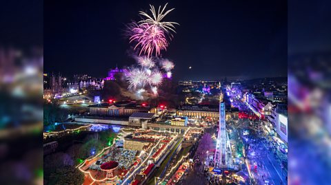 Hogmanay celebrations in Edinburgh on New Years Eve. The city is full of colourful lights and markets from the festive period and celebrations and in the background, bright pink and purple fireworks can be seen in the distance