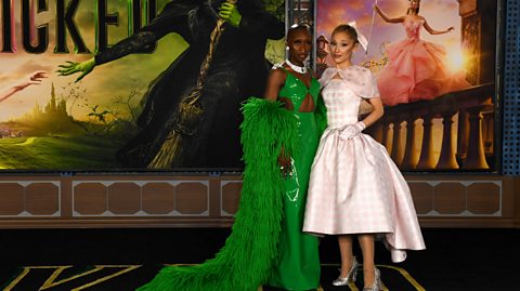 Cynthia Erivo left, wears a shiny, long green dress with cut outs and a fluffy green coat, which have fallen off the shoulder. Next to her on the right, Ariana Grande wears her blonde hair in a bun with a big light pink bow, which matches her gingham pink baby doll dress with puffy sleeves. They are at a Wicked premiere with film posters on the wall behind them