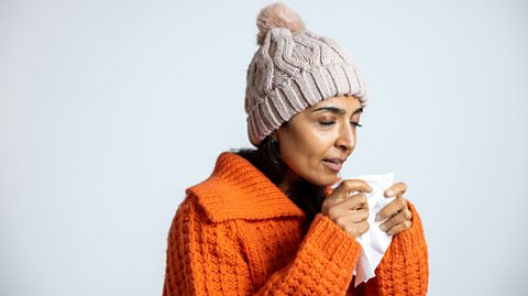 Person wearing a thick orange knitted sweater and a beige cable-knit winter hat, holding tissues against a light grey background.