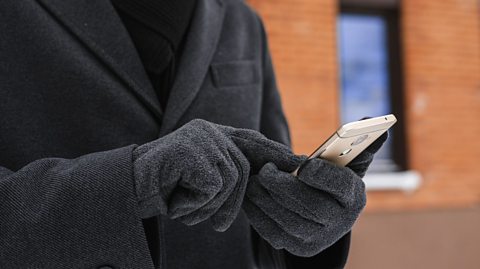Person wearing a dark winter coat and gloves using a smartphone outdoors in front of a brick building