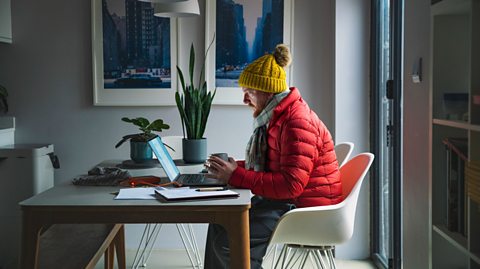 Person in a red puffer jacket and yellow knit hat working on a laptop at a modern dining table with papers, a coffee mug, and framed cityscape photos on the wall.