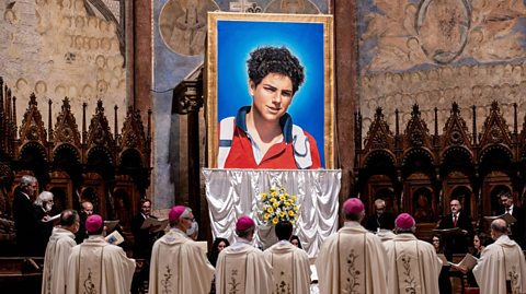 The beatification of Carlo Acutis. Pictured, a large photo of Saint Carlo Acutus, wearing a red jacket and a backpack against a blue background, surrounded by members of the Vatican in traditional attire of cream robes and red skullcaps