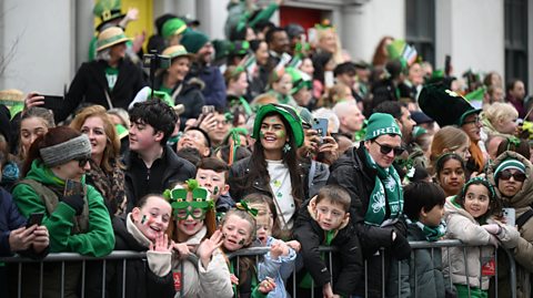 Behind a barrier, crowds of adults and children smile during the Dublin St Patrick's Day parade, wearing green and Irish themed hats, scarfs, glasses. Some have shamrocks - a three-leaf clover - on their faces