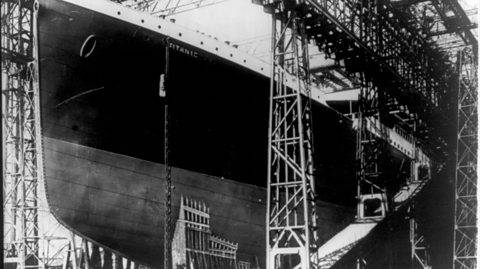 A black and white photo of a large ship, The Titanic, surrounded by scaffolding as it sits in a shipyard