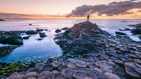 A tourist stands on the stone at the Giant's Causeway, Northern Ireland, looking out at the water as the sun sets