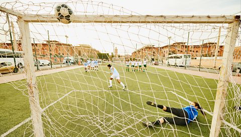 A wide shot taken from behind a goal through the nets shows a footballer player kicking a ball into the net during a penalty kick. The two teams are in the background lined up watching on the grass and the goalkeeper is on the floor after missing the ball