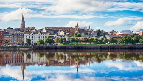 The city of Derry/Londonderry in Northern Ireland. On a sunny day, the city is reflected in the water and there are trees, buildings and churches on the city skyline