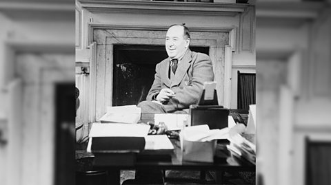 A black and white photo of author C S Lewis wearing a suit and tie, smiling. He sits behind his desk which is full of files and in front of a large, marble fireplace