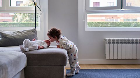 Toddler in polka-dot pyjamas leaning toward a baby lying on a grey sofa in a bright living room with large windows and modern decor