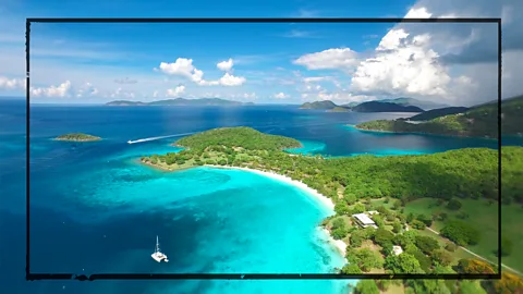 Getty Images A lush green Caribbean island is shown against blue skies and surrounded by blue water. A boat is seen in the foreground and the skies are full of fluffy white clouds. A stylised black border has been edited on top. (Credit: Getty Images)