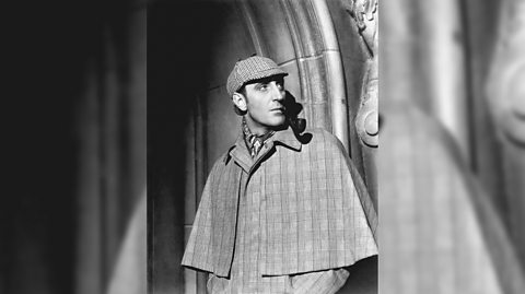 A black and white photo of actor Basil Rathbone as Sherlock Holmes, wearing an Ulster overcoat which is tweed and with a cape and a flat cap as he leans against an archway