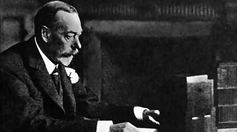 A black and white photo of King George V, wearing a suit and sitting at a desk, giving the 1934 Royal Christmas message by radio