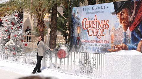 Actor and comedian Jim Carrey, wears black trousers, a black shirt, a grey blazer and sunglasses at the 62nd Cannes Film Festival for Disney's A Christmas Carol. He leans against a barrier, smiling and looking up the the sky. Behind the barrier is a large poster for the animated film, showing Scrooge, a Victorian character holding a candle, wearing a hat, scarf and small glasses, looking unpleasant. In front of the poster is a manmade winter wonderland, with snow dusted on the floor, a Christmas tree with red baubles and a large silver bauble and large red bauble