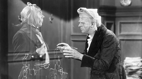 The ghost of Jacob Marley, on the left, is transparent tied in thick, metal chains. He wears a Victorian suit with bandages around his head and speaks to Ebenezer Scrooge on the right. Scrooge wears a sleeping hat and robe and looks fascinated by the ghost