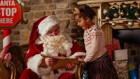 Father Christmas, with a white beard and glasses, wears a red hat and suit which has white fur lining. He sits by a fireplace decorated with baubles and reads his naughty and nice list. Next to him stands a young girl, wearing a reindeer ear headband, her hair in pigtails, a festive jumper and metallic skirt. She smiles as she spots her name on the nice list and leans against the arm of the chair