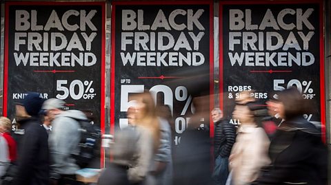 Shoppers walk past a shop window in a blur, big black, white and red signs declare 50% savings for Black Friday weekend