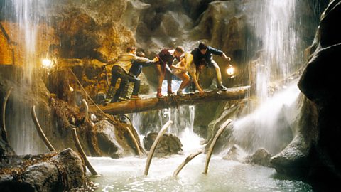 A still from the movie The Goonies. Several characters are walking across a log within a cave, with waterfalls either side of them and water below the log