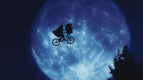 A still from the movie E.T. the Extra Terrestrial. Elliott rides his bike through the sky, with E.T. in his bike's basket, against the backdrop of the moon