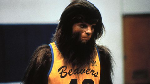 A still from the movie Teen Wolf. Michael J. Fox's character is in werewolf mode, wearing a yellow basketball jersey with blue trim and the name Beavers on the front
