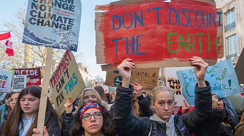 A crowd of students and young people hold placards one home made red background says 'Don't discount the earth', another printed one calls for System Change not climate change