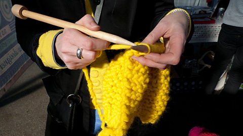A women uses large wooden needles to knit a bright yellow garment, maybe a scarf