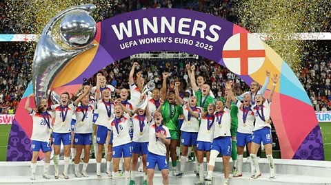 The England women's football team lift the European Championships trophy on stage, with a winners banner behind them. Gold glitter is being shot into the air.