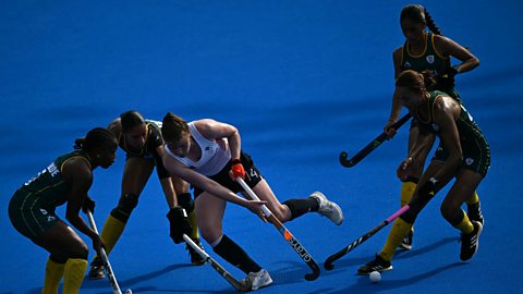 Tess Howard, wearing a white vest and navy shorts, is surrounded by four South African players in dark green and yellow. All are holding hockey sticks on a blue astroturf pitch