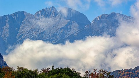 Grey mountains are slightly covered by white clouds. The sky is bright blue with a few soft clouds, and there are green trees at the bottom.