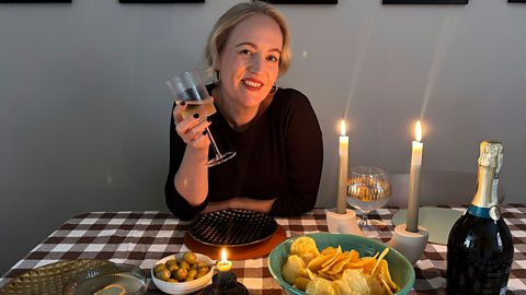 Lauren sitting at a dinner table with crisps and olives in front of her and two lit candles and a bottle of wine on the table too