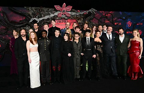 The cast of Stranger Things attend the world premiere of season 5 in Los Angeles. Behind them is a large wall which is red and has lots of black, scaly tentacles on it