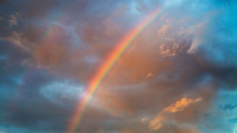 Vibrant rainbow arching across a dramatic sky with blue tones and orange-tinged clouds