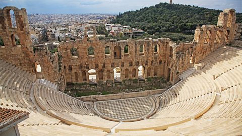 An ancient Greek theatre. There are ruins behind the stage and lots of tiered seats. 