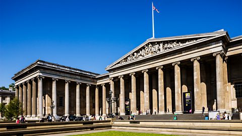 A huge stone building with tall columns in front. There are carvings of people and shapes on the triangular roof section.