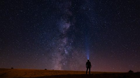 Milky Way stretches across the night sky, which is dark blue with hundreds of bright stars as small as dots in the distance. A silhouette of a man looks up at the sky, wearing a helmet with a bright torch attached