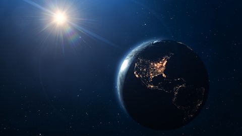 An image of space, with the bright, yellow sun and on the far left beaming light onto Earth, which is mainly dark, with the Americas on the night side of the planet