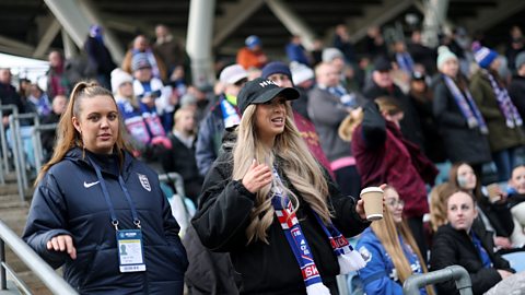 GK Barry, wearing a black coat and black baseball cap, with a blue, red and white Ipswich Town scarf, walks down steps in a football stadium, holding a brown coffee cup