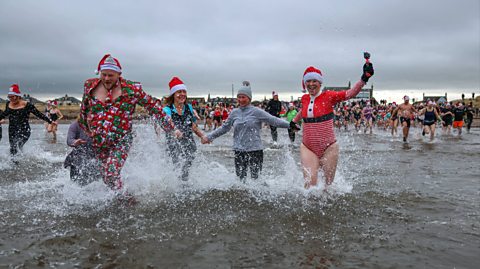 On a dull Boxing Day, crowds run into the water in Ayr, Scotland for the annual dip. Image shows four people at the forefront, a man wearing a green and red Christmas suit and matching hat crashing into the water, next to three women also running into the cold water holding hands. From the left, the first woman has a red and white Santa hat on, a blue top and black pinafore, next to her another lady has black leggings, a grey jumper and matching wool bobble hat on and to the far right, the third woman has a red and white Santa hat on with black gloves and a red swimming costume with a Santa style cardigan over it
