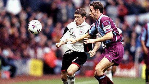 Gareth Southgate in a claret and blue shirt, claret shorts and socks competes for the ball with a Darlington player, in white and black