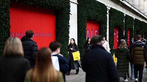 Shoppers on a busy London street are carrying bright yellow shopping bags full of items they bought in the Boxing Day sales. On this street, a department store has windows covered in red signs and bold writing saying 'Sale.' The border of the windows are full of festive foliage