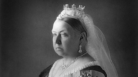 A black and white image of Monarch Queen Victoria, with a stern expression, wearing a crown and white veil, pearl drop earrings, a necklace and a ruffled dress