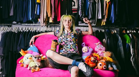 Emma Rogue sits on a bright pink huge chair surrounded by retro plushies and clothing on big rails behind her