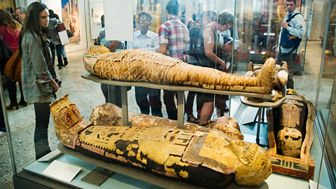 A photo of the Egyptology room in the British Museum with visitors looking at mummies and sarcophagus's.