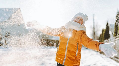 A road full of houses and trees covered in snow. At the forefront of the image is a young toddler boy in a bright yellow waterproof coat, with a white hat and matching scarf, closing his eyes as he dusts snow off his gloves