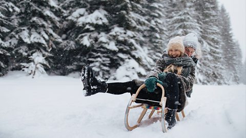 A young couple, surrounded by snow and trees, sledding. They both wear winter hats and chunky knitted scarves