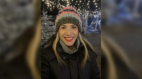 BBC senior weather presenter Kay Crewdson wears a multicoloured knitted hat and black coat as she smiles for the camera. Behind the selfie is festive lights and a Christmas tree